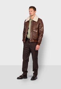 Hnědá kožená bunda s krémovým ovčími límcem, se předními kapsami, spárovaná se zelenou košilí, tmavě hnědými kalhotami a černými botami.