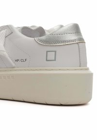 Zapatilla blanca con detalles plateados, parte superior de cuero suave, cuello acolchado y suela de goma gruesa y texturizada. Presenta un logotipo mínimo.