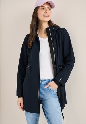 Femme portant une veste à capuche bleu marine par-dessus une chemise blanche et un jean bleu clair, une main dans la poche et une casquette rose clair.