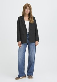 Črn blazer z ožjim ovratnikom, enojnim gumbom in sprednjimi žepi, kombiniran z ohlapnimi modrimi kavbojkami in rjavimi športnimi copati.