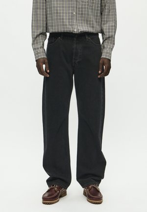 Bredde ben sorte denim jeans med en struktureret finish. Kombineret med en ternet skjorte med knapper og brune lædersko med dekorative snørebånd.