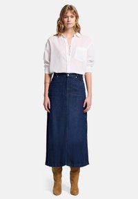 Gonna maxi in denim blu scuro con vita aderente e silhouette dritta, abbinata a una camicia bianca a maniche lunghe e stivaletti beige.