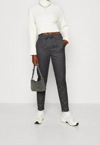 Hoch geschnittene, grau gestreifte Hose mit einem Kordelzugbund, kombiniert mit einem weißen, gerippten Crop-Sweater und einer grauen Handtasche. Weiße Sneakers runden den Look ab.