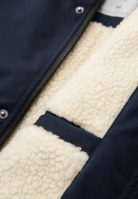 Veste en tissu extérieur bleu marine avec une doublure douce en fausse peau de mouton crème et une poche intérieure bordée d'un passepoil assorti bleu marine. La fermeture éclair est visible.