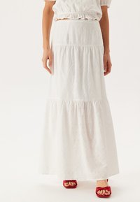 Jupe maxi blanche à plusieurs niveaux en coton brodé, ornée de motifs floraux découpés. Complétée par des sandales à talons rouges.