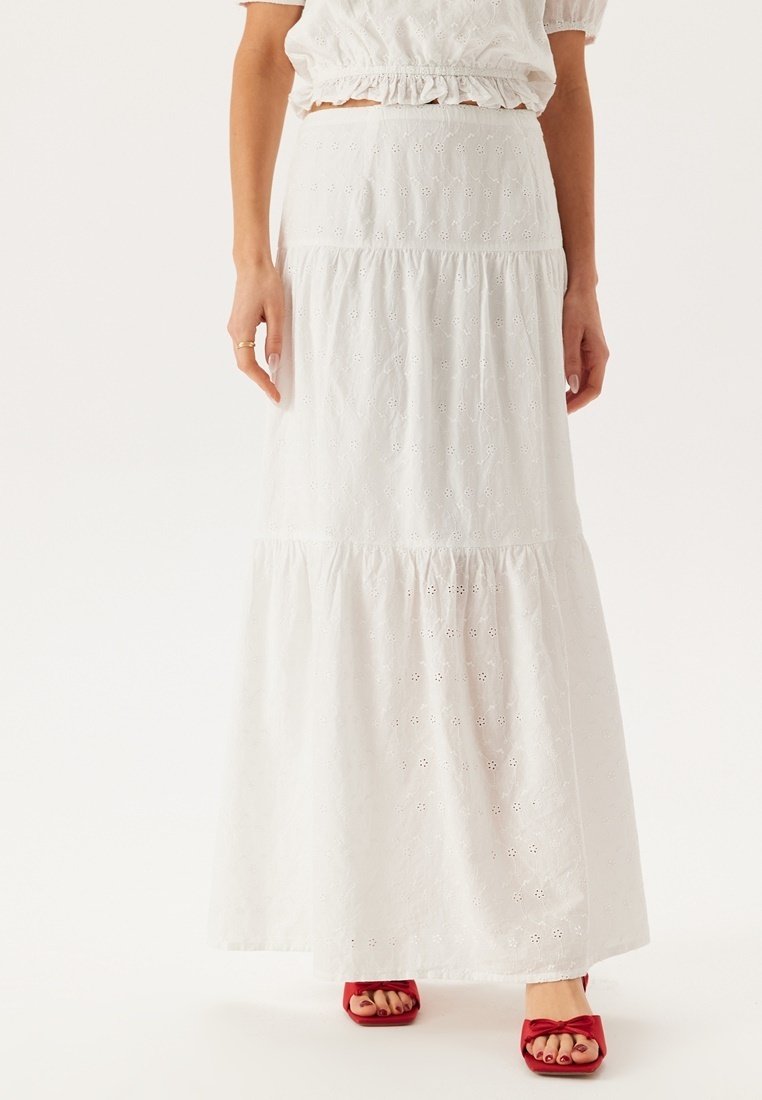 Jupe maxi blanche à plusieurs niveaux en coton brodé, ornée de motifs floraux découpés. Complétée par des sandales à talons rouges.