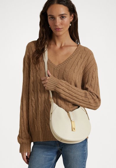 Geantă de umăr din piele albă cu o formă curbată, accesorii din aur și un finisaj texturat, purtată peste un pulover maro cu tricotaj de tip cable-knit.