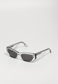 Lunettes de soleil grises et angulaires avec une monture transparente, des verres sombres et des accents noirs mis en évidence sur les branches. Nom de la marque visible sur le côté.
