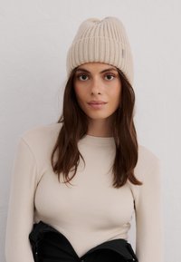 Gorro de punto acanalado beige con la parte superior redondeada y una pequeña etiqueta de logo. El modelo lleva una camiseta de manga larga de color crema, con detalles de tejido texturizado.