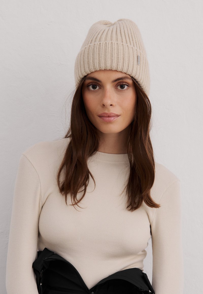 Gorro de punto acanalado beige con la parte superior redondeada y una pequeña etiqueta de logo. El modelo lleva una camiseta de manga larga de color crema, con detalles de tejido texturizado.