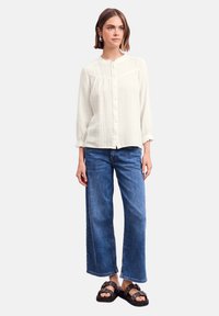Blusa bianca a trama con colletto alla cinese e chiusura a bottoni, abbinata a jeans blu a gamba larga e sandali neri con accessori decorativi.