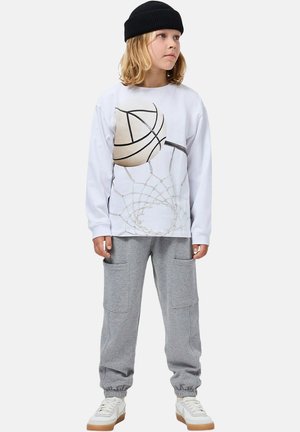Camicia bianca a maniche lunghe con grafica da basket, pantaloni della tuta grigi con tasche laterali e sneakers bianche con suole color cuoio. Berretto nero in testa.