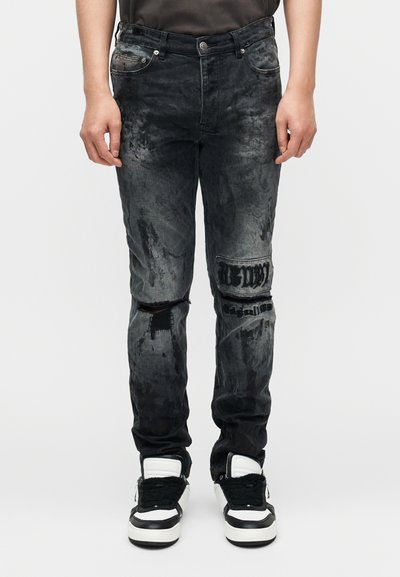 Μαύρα τζιν denim με σκισμένες περιοχές, ξεθωριασμένο φινίρισμα και κεντημένες λεπτομέρειες στο γόνατο. Φορεμένα με μαύρα και λευκά αθλητικά παπούτσια.