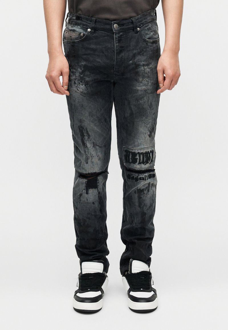 Μαύρα τζιν denim με σκισμένες περιοχές, ξεθωριασμένο φινίρισμα και κεντημένες λεπτομέρειες στο γόνατο. Φορεμένα με μαύρα και λευκά αθλητικά παπούτσια.