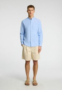 Svijetloplava košulja s ovratnikom u obliku trake, uparena s bež šortsevima i smeškim natikačama. Ležerna ljetna odjeća.