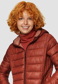 Czerwona kurtka puchowa z pionowym pikowaniem, zamek błyskawiczny i kaptur, noszona na brązowej bluzie z długim rękawem. Miękki, błyszczący materiał.