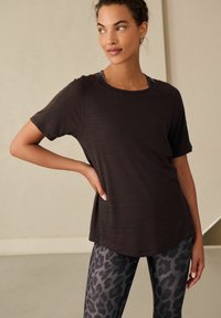 Donkerbruin sporttop met korte mouwen en een gestructureerde afwerking, gecombineerd met zwarte en grijze legging met luipaardprint. Relaxte pasvorm en ronde hals.