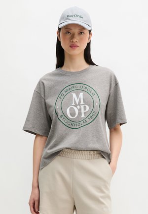 Γυναίκα που φοράει γκρι μπλουζάκι με το λογότυπο Marc O’Polo, μπεζ σορτς με ελαστική μέση και λευκό καπέλο Marc O’Polo, στέκεται με το ένα χέρι στην τσέπη.