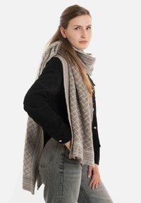 Beige sjaal met een diamantpatroon, losjes om de nek gedragen, in combinatie met een zwarte ribgebreide sweater en lichtgrijze jeans.