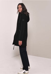 Manteau en polaire noir avec une capuche, une fermeture éclair à l'avant et un cordon de serrage. Présente un badge sur le côté, stylé avec un pantalon sombre et des baskets blanches.