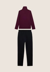 Maglione a collo alto lungo di colore viola scuro abbinato a pantaloni neri con vita elastica, stesi su uno sfondo beige.