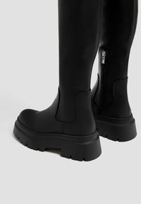 Bottes noires cuissardes avec une texture lisse, des semelles épaisses en caoutchouc et une fermeture éclair latérale. Design élégant avec un bout arrondi et une construction robuste.
