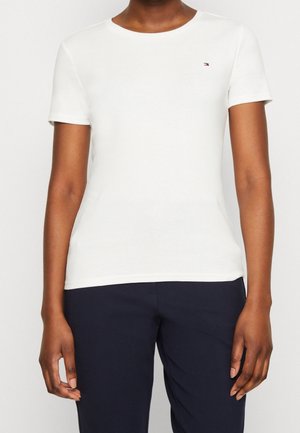 Osoba ubrana w białą, gładką koszulkę z krótkim rękawem z małym logo na lewej piersi oraz ciemnogranatowe spodnie, stojąca na jednolitym tle.