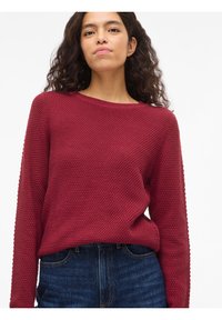 Νεαρή γυναίκα με σγουρά μαλλιά, ντυμένη με μπλούζα υφής σε σκούρο κόκκινο χρώμα και μπλε τζιν παντελόνι, σε λευκό φόντο.