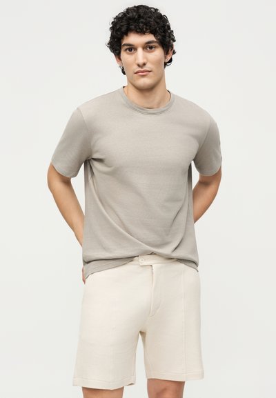 Νέος άνδρας με σγουρά μαύρα μαλλιά που φοράει ένα ανοιχτό γκρι κοντομάνικο T-shirt και εκρού κοντά παντελόνια με υφή, στέκεται μπροστά σε απλό φόντο.