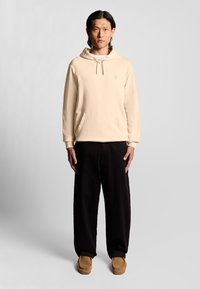 Beige Kapuzenpullover mit einer Fronttasche und Logo, kombiniert mit schwarzen weitem Hosen und braunen Slippern. Glatte Stoffstruktur.