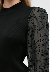 Schwarzer Rippenpullover mit hohem Kragen, ausgestattet mit schwarzen, transparenten Puffärmeln, die mit floralen Stickereien verziert sind.
