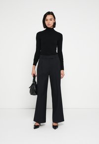 Sort rullekravebluse kombineret med sorte pinstripe bukser med høj talje og vide ben. Modellen holder en sort håndtaske med struktur. Spidse sorte sko.