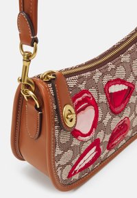 Brązowa torba na ramię z tkaniny z czerwonymi akcentami w kształcie ust, złotym okuciem i brązowym skórzanym paskiem. Posiada zamek błyskawiczny na górze.