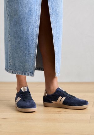Jambes portant une jupe en jean bleu clair avec une fente à l'arrière et des baskets en daim bleu marine avec des accents beige, debout sur un sol en bois.