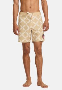 Beige swim shorts with a white geometric pattern, drawstring waist, and a red patch logo on the left leg. Smooth texture, mid-thigh length.