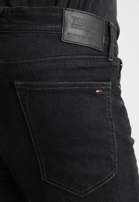 Jeans de mezclilla negra con una etiqueta de cintura de cuero texturizado que dice "TOMMY JEANS." Presenta costuras en contraste y un pequeño acento de una bandera en el bolsillo.