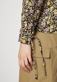 Schwarzes, blumiges Langarm-Oberteil mit gelben, lila und grünen Blumen, mit einer Schleifendetail am Bündchen, getragen mit einer beige Hose mit Taschen.
