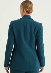 Tealfärgad figursydd kavaj med slagen krage, mjuk textur, strukturerade axlar och en enda ryggslits. Har minimala design detaljer.