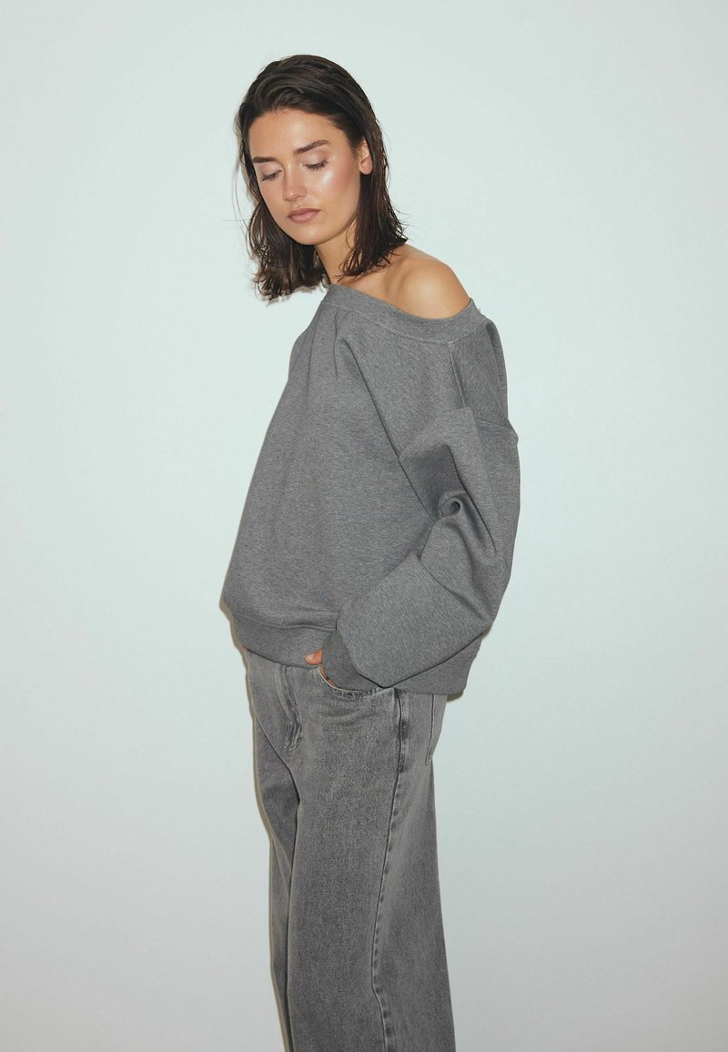 Grijze oversized sweatshirt met een off-shoulder ontwerp. Voorzien van brede mouwen en geribbelde boorden. Gecombineerd met losse grijze jeans. Eenvoudige, neutrale kleuren.