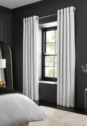 Tende bianche a tutta altezza appese a una bacchetta di metallo intorno a una finestra con cornice nera in una camera da letto grigio scuro con un letto bianco e un tappeto beige.