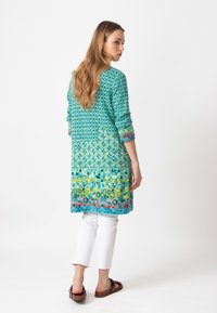 Pitkä turkoosi tunika, jossa on geometrisia kuvioita, väri muuttuu asteittain keltaiseksi ja hemman koristeena on kukka-aiheita, yhdistetty valkoisiin housuihin.