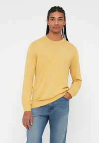 Gul strikkegenser med lange ermer og rund hals, kombinert med lyseblå jeans. Genseren har en myk tekstur og ribbede mansjetter.