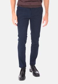 Uomo che indossa pantaloni in velluto a coste navy slim-fit e scarpe stringate in pelle nera, in piedi su uno sfondo bianco.