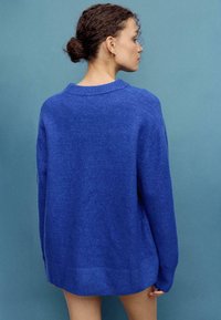 Modro velik oprijet pulover z rebrastim ovratnikom in manšetami, z mehko teksturo in ravnim robom, prikazan od zadaj.