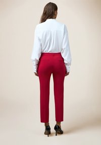Blouse blanche à manches longues associée à un pantalon rouge sur mesure. Le pantalon présente une coupe slim avec des poches, rehaussé par des chaussures à talons noires.