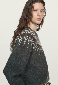 Jeune femme aux longs cheveux ondulés portant un pull en tricot gris foncé avec des motifs géométriques blancs et marron sur les épaules.