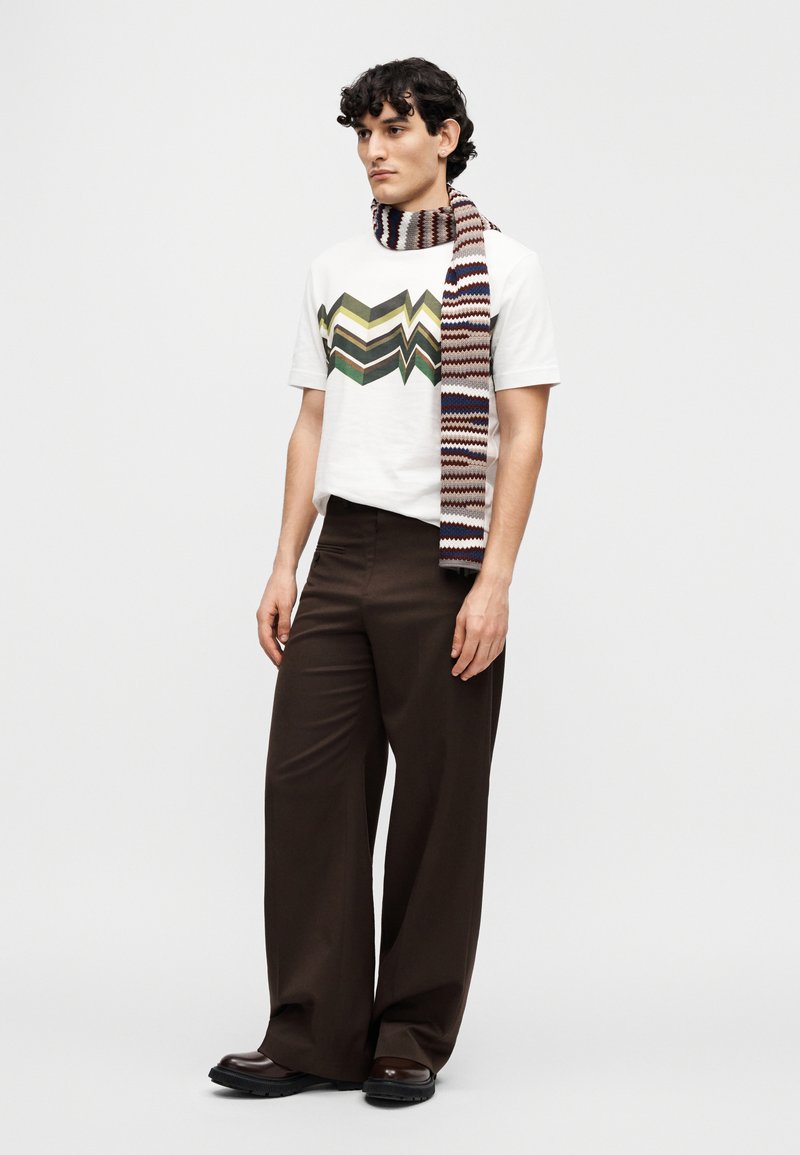 White cotton t-shirt with zigzag green, brown, and blue graphic. Brown wide-leg trousers; knitted scarf with red, white, and blue pattern.