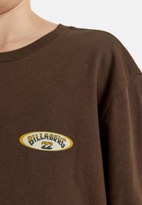 Brązowa koszulka bawełniana z okrągłym dekoltem, z białą i żółtą naszywką z logo, która wyraźnie informuje "BILLABONG" na piersi.