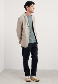 Beige linnblazer över en grön skjorta, kombinerad med navybyxor och flerfärgade sneakers. Har en framficka och en slät textur.