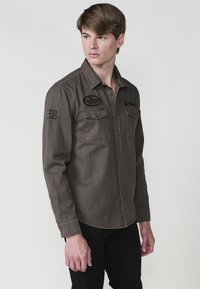 Donkergrijze langsliever shirt met twee borstzakken, knoopsluiting en geborduurde patches op de schouders. Gemaakt van een gladde stof.